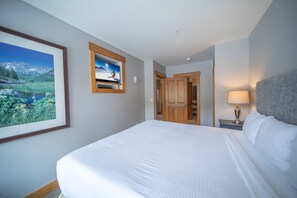Una habitación de hotel con una cama grande, un cuadro enmarcado con un paisaje de montaña, una puerta de madera y una mesita de noche con una lámpara.