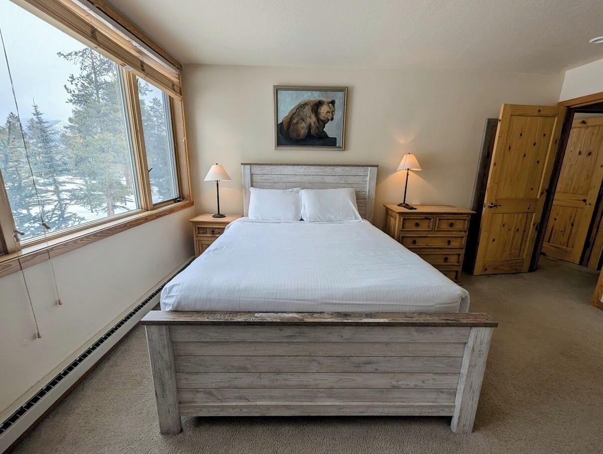 A bedroom with a wooden bed, a painting of a bear on the wall, and a view of trees through the window.