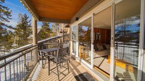 Une terrasse en bois avec une table et des chaises, une porte vitrée donnant sur un espace intérieur avec un canapé, et une vue sur des arbres et un bâtiment au loin.