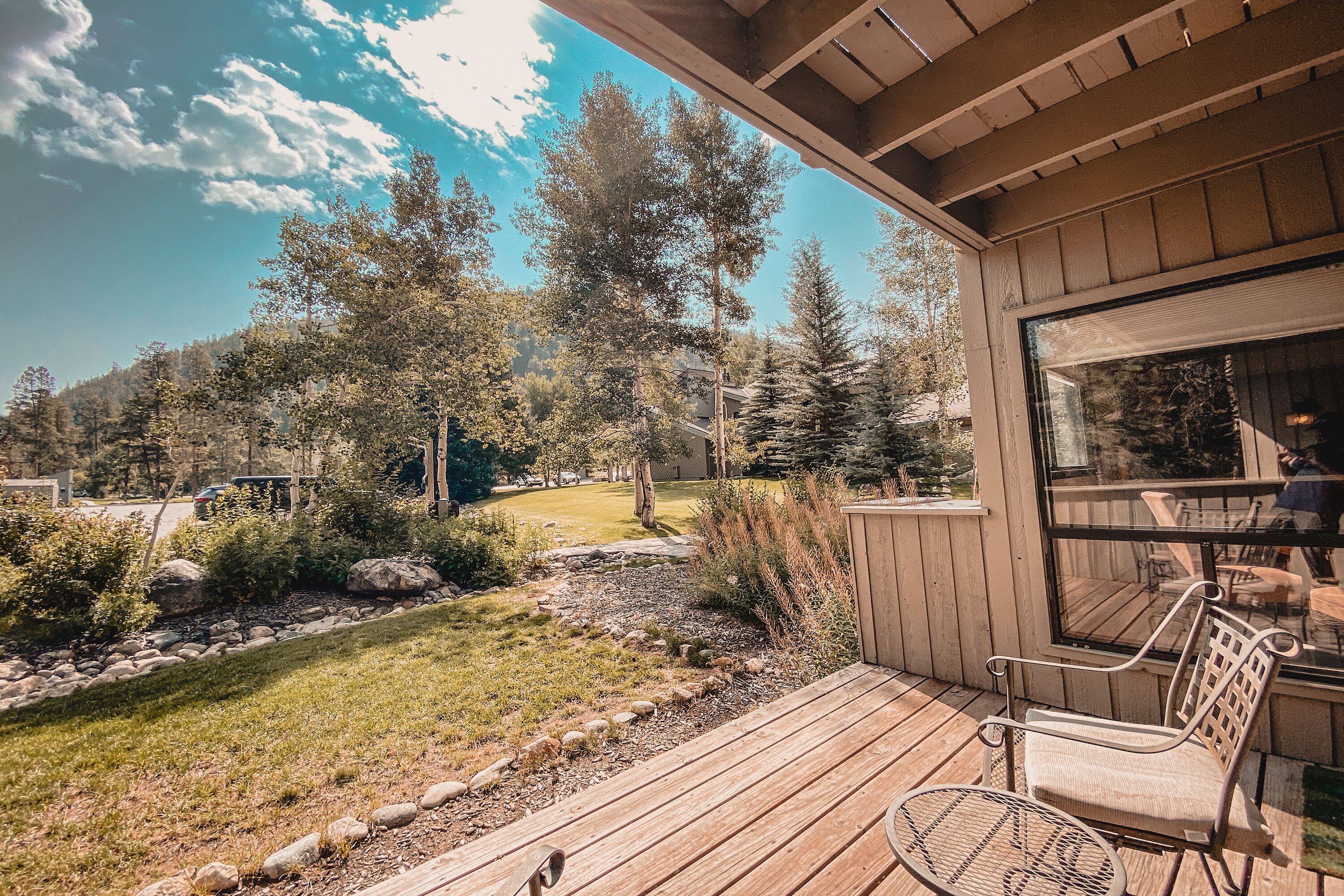 A wooden deck with a metal chair and table, overlooking a landscaped garden with trees and a clear sky.