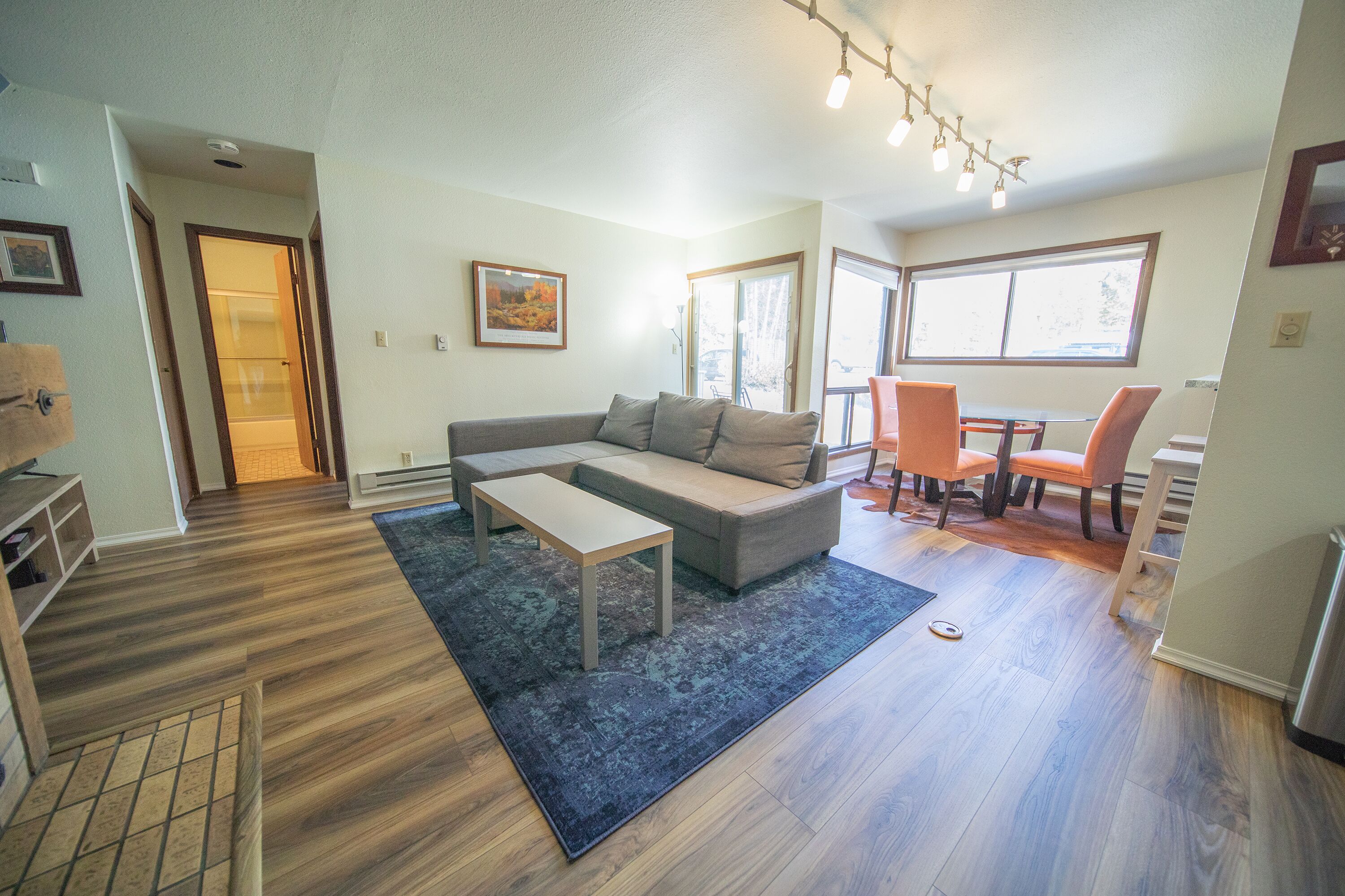 A modern living room with a sofa, coffee table, and chairs. There is a dining area with a table and chairs. The room has hardwood flooring and large windows.