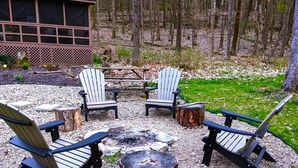 A fire pit area with Adirondack chairs and a table, surrounded by a gravel ground with trees in the background.