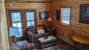 A cozy cabin interior with a sofa, armchair, and a small dining table. There are two windows with blinds, a lamp, and a framed picture on the wall.