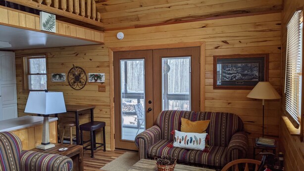 A cozy cabin interior with a wooden floor, a striped sofa, a dining table with chairs, and a window with blinds.