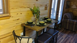A wooden interior with a dining table set for four, chairs, a vase with flowers, and a window with blinds.