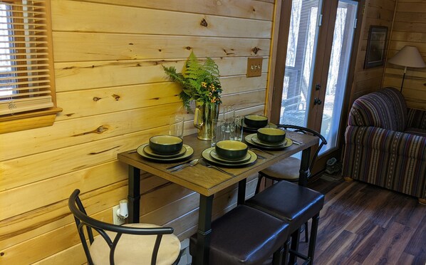 A wooden interior with a dining table set for four, chairs, a vase with flowers, and a window with blinds.