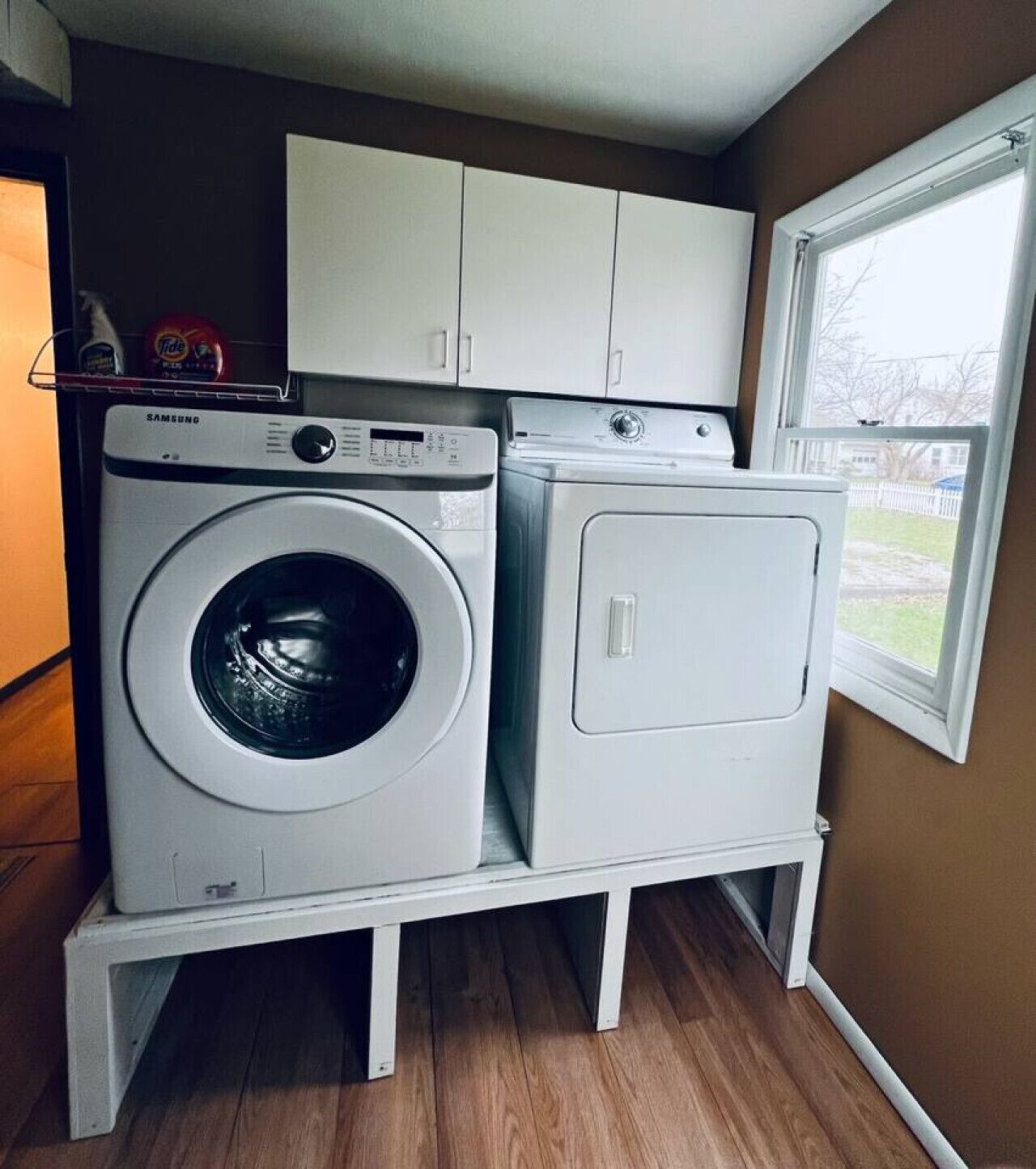 Un espace buanderie compact comprenant un lave-linge et un sèche-linge à chargement frontal, des armoires blanches au-dessus et une fenêtre sur le côté droit.