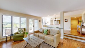 Ein geräumiges Wohnzimmer mit Sofa, Sessel und Couchtisch. Große Fenster mit Jalousien, eine Küchenzeile und eine Treppe, die zu einer weiteren Ebene führen.
