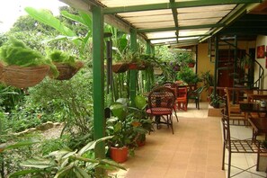 A covered patio area with hanging planters and potted plants, wooden furniture, and a tiled floor. - Hotel Los Jardines de Monteverde (Monteverde)