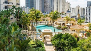 A resort with a central swimming pool surrounded by palm trees and a view of high-rise buildings in the background.