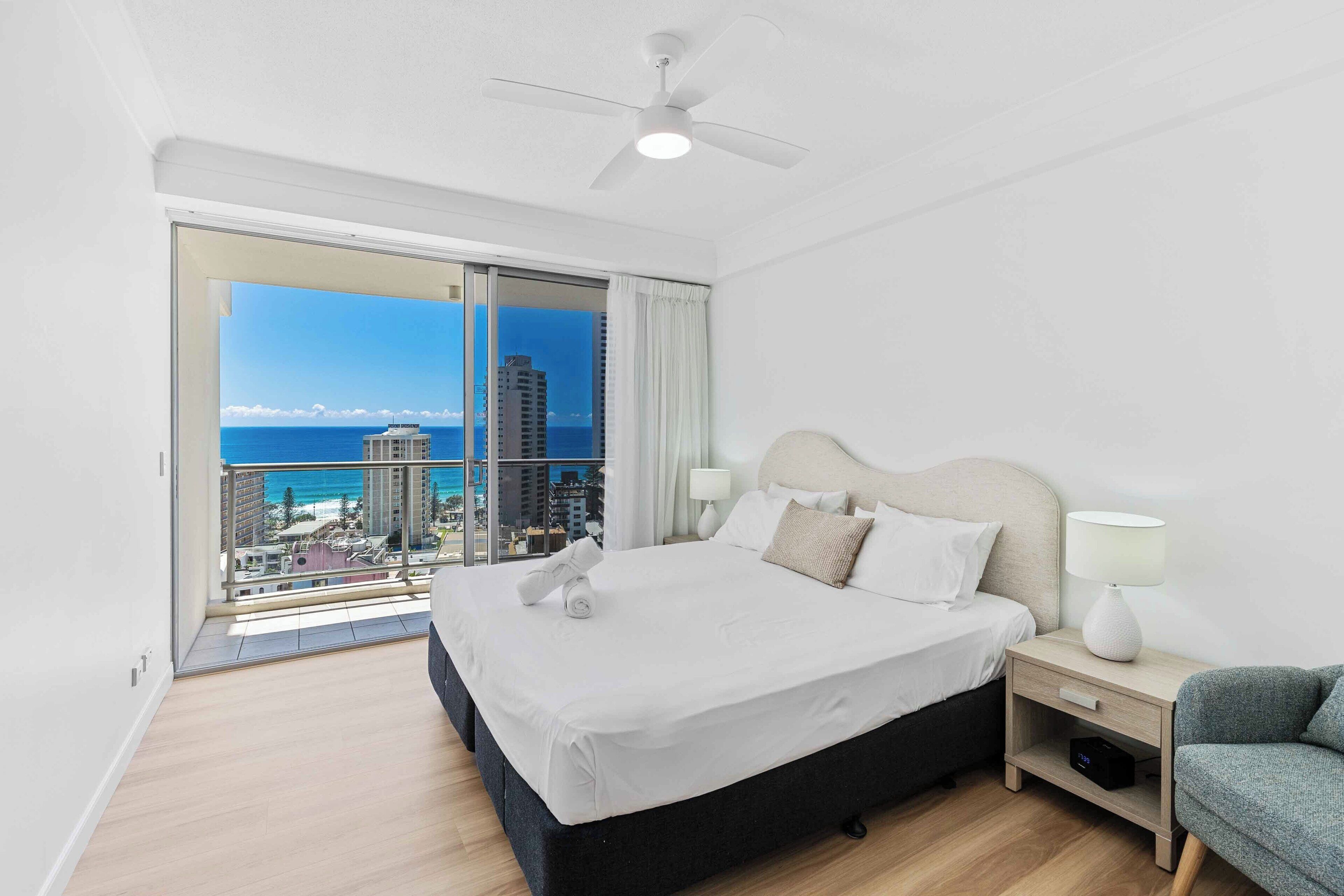 A modern hotel room with a large bed, a nightstand, a lamp, and a view of the ocean and cityscape.