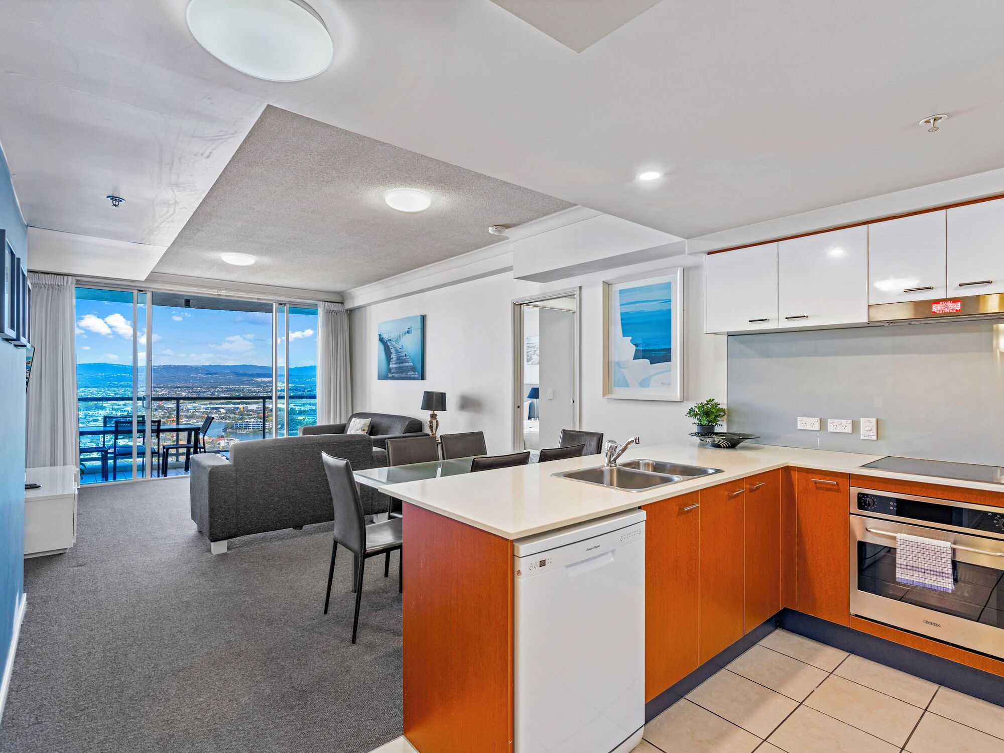 A modern kitchen with a dishwasher, oven, and sink. There is a dining area with a table and chairs, and a view of the city through large windows.