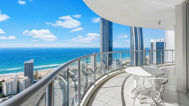 A balcony with a curved glass railing overlooking a beach and ocean, featuring a white table and two chairs.