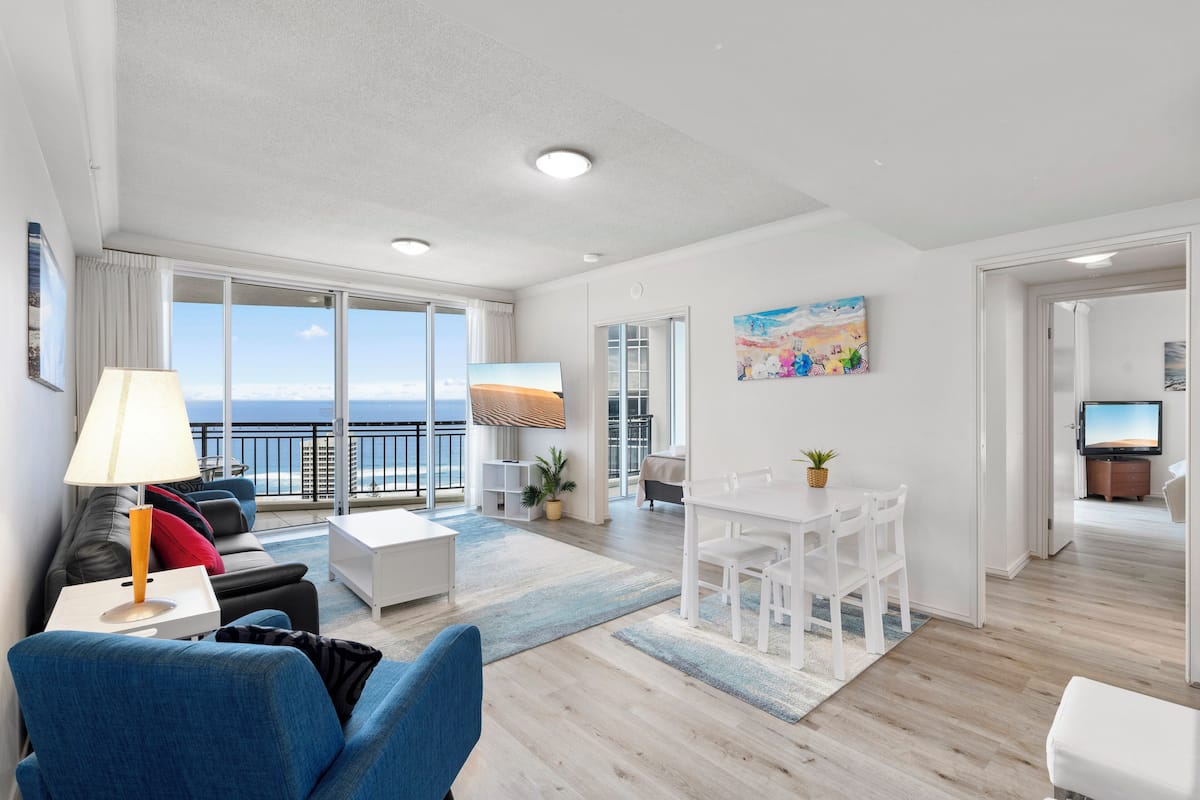 Ett modernt vardagsrum med en soffa, ett matbord och en tv. Det finns en balkong med utsikt över havet.