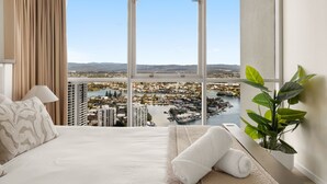 A hotel room with a large window offering a city view, a bed with white linens, a bedside plant, and a lamp.