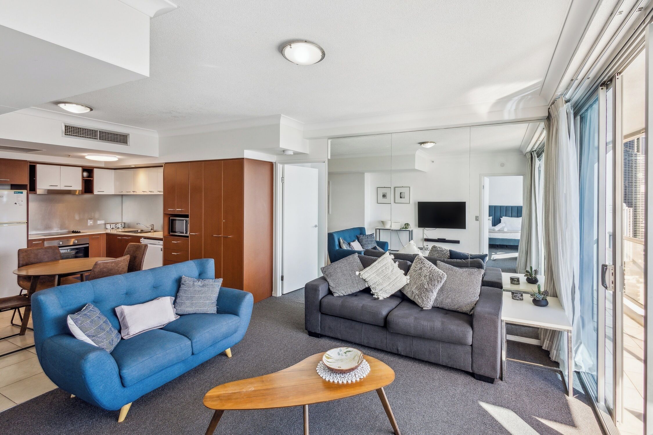 A modern living room with a blue sofa, a grey sofa, and a wooden coffee table. There is a kitchen area with wooden cabinets and a dining table.