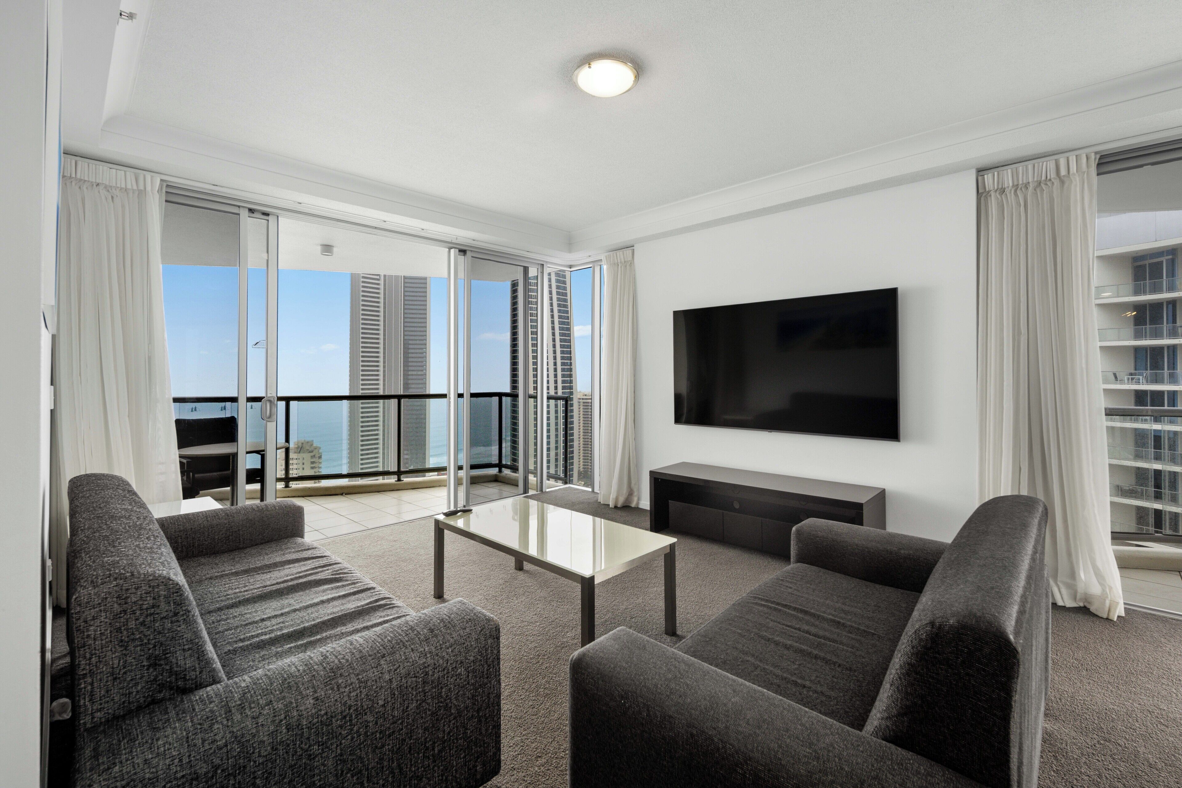 A modern living room with a flat-screen TV, grey sofas, and a glass coffee table.
