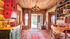 A rustic cabin interior with wooden floors, a red wall with stars, a chandelier, and a large window. - Log Cabin at Old Glory Ranch | Take a trip back in time! (Wimberley)
