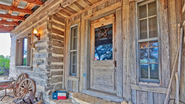 A rustic log cabin with a stone foundation, wooden exterior, and a porch with a bucket of firewood. - Log Cabin at Old Glory Ranch | Take a trip back in time! (Wimberley)