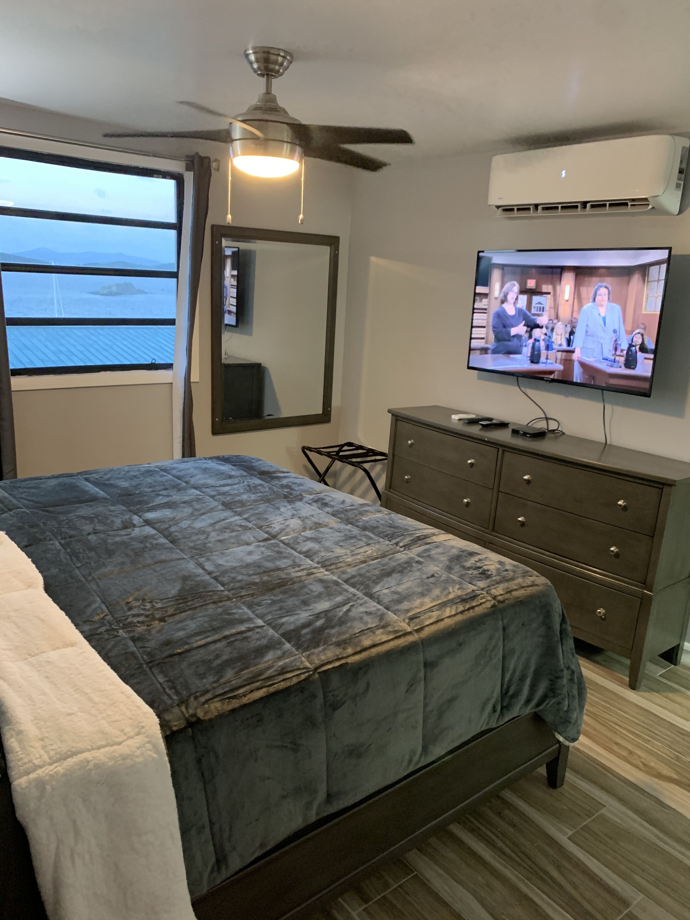 Een slaapkamer met een bed, een aan de muur bevestigde televisie, een plafondventilator en een raam met uitzicht op de oceaan.