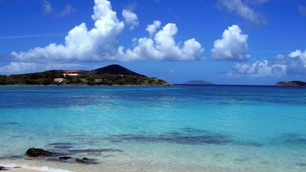 Una playa con agua turquesa cristalina, una orilla arenosa y una pequeña isla con edificios al fondo.