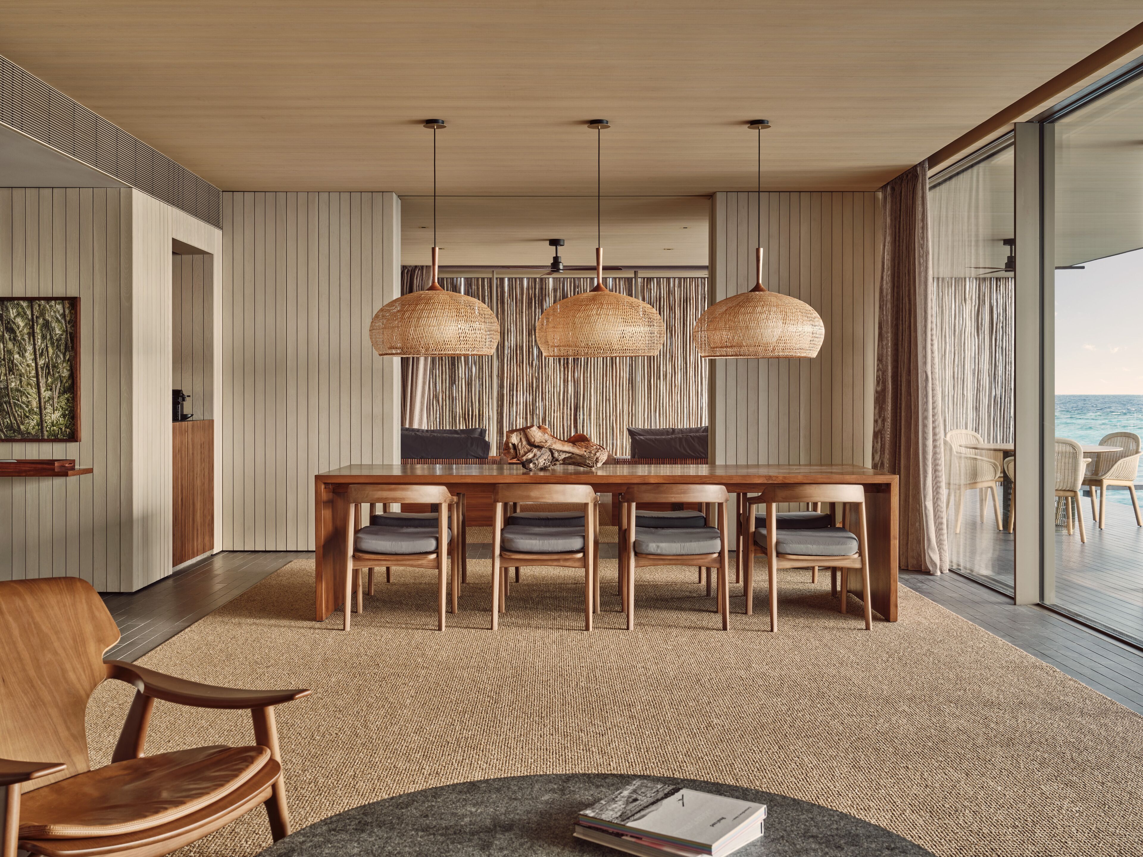 Un espace de repas moderne avec une table et des chaises en bois, des suspensions lumineuses en fibres tressées et une vue sur l’océan grâce à de grandes fenêtres.
