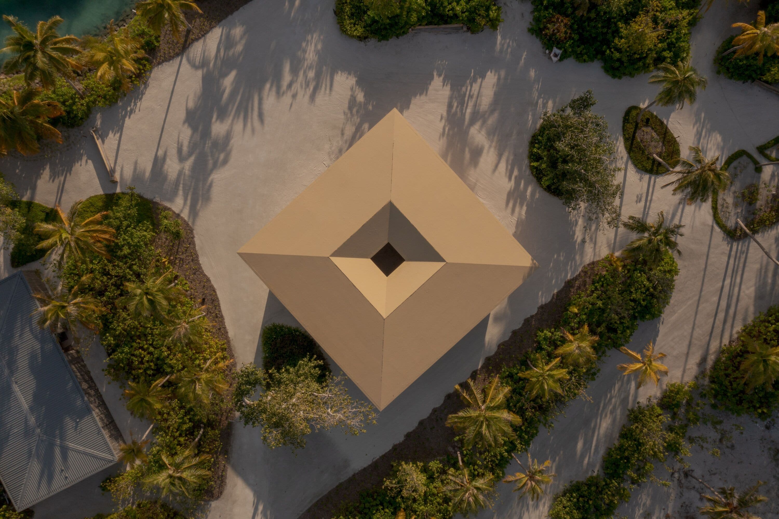 An aerial view of a resort with a central geometric structure surrounded by palm trees and a sandy beach.
