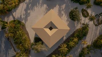 An aerial view of a resort with a central geometric structure surrounded by palm trees and a sandy beach.