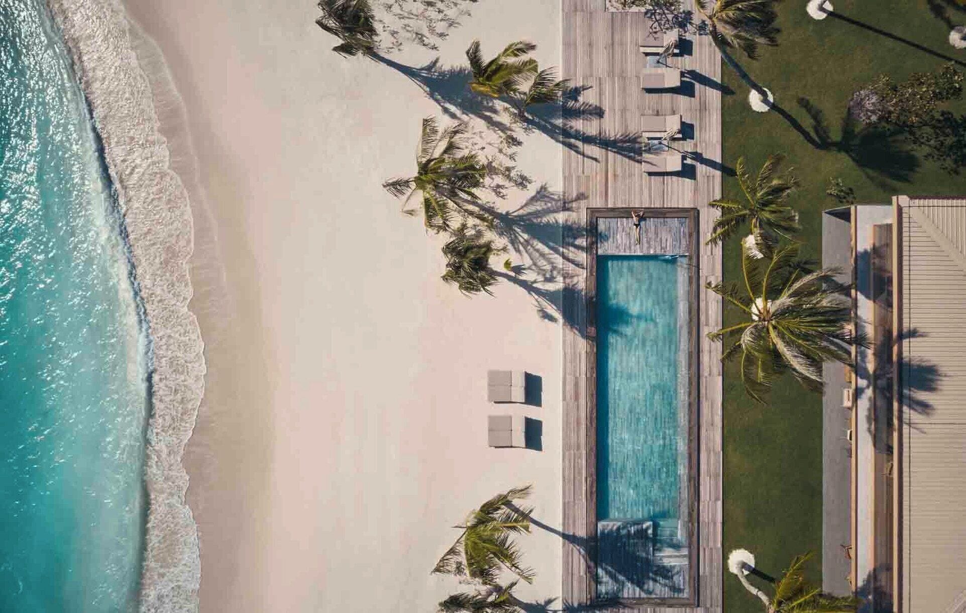 An aerial view of a beach resort with a swimming pool, palm trees, and a sandy beach.