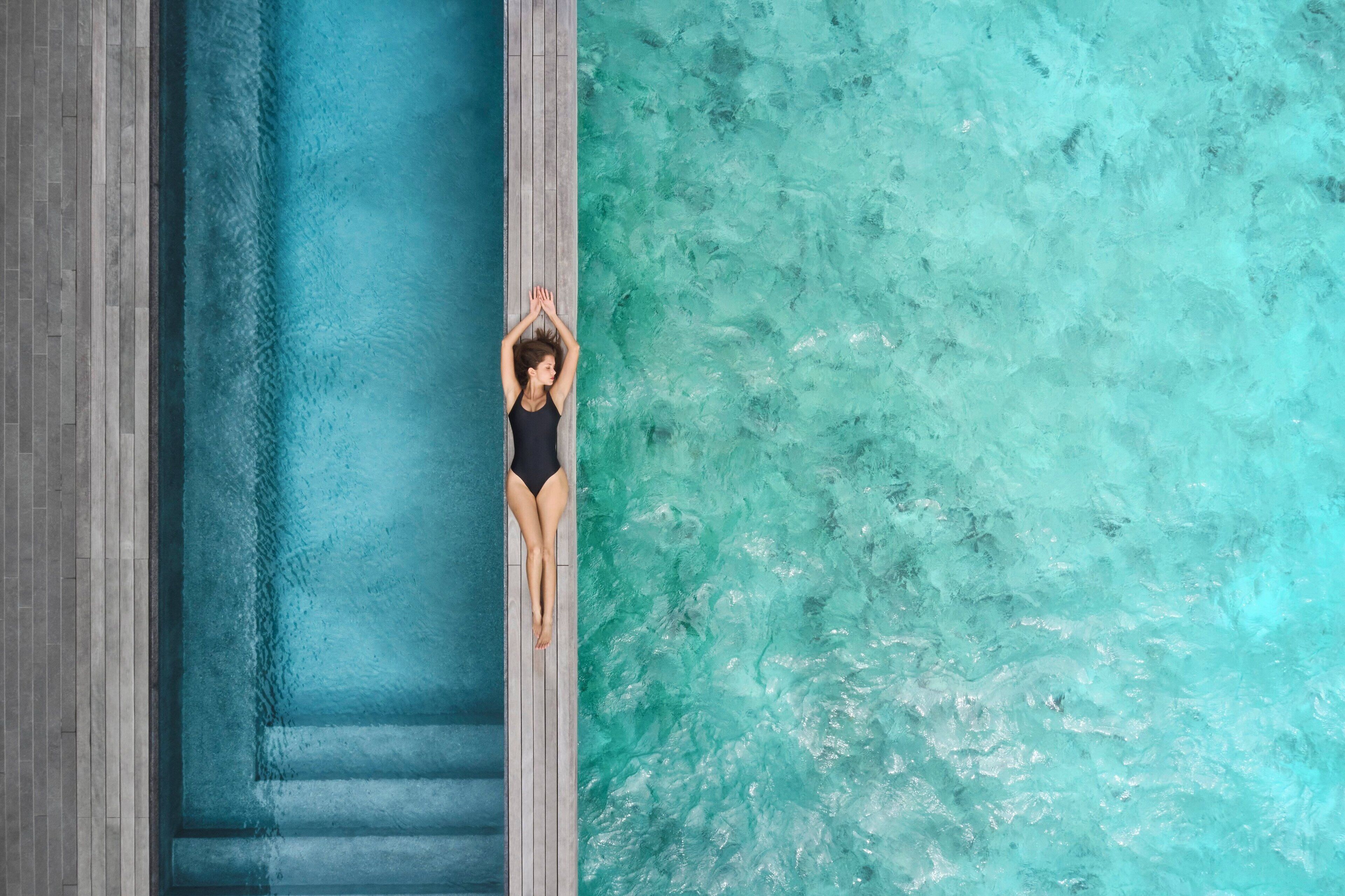 Une personne vêtue d’un maillot de bain noir se tient debout dans une piscine, avec une terrasse en bois d’un côté et une rive rocheuse de l’autre.