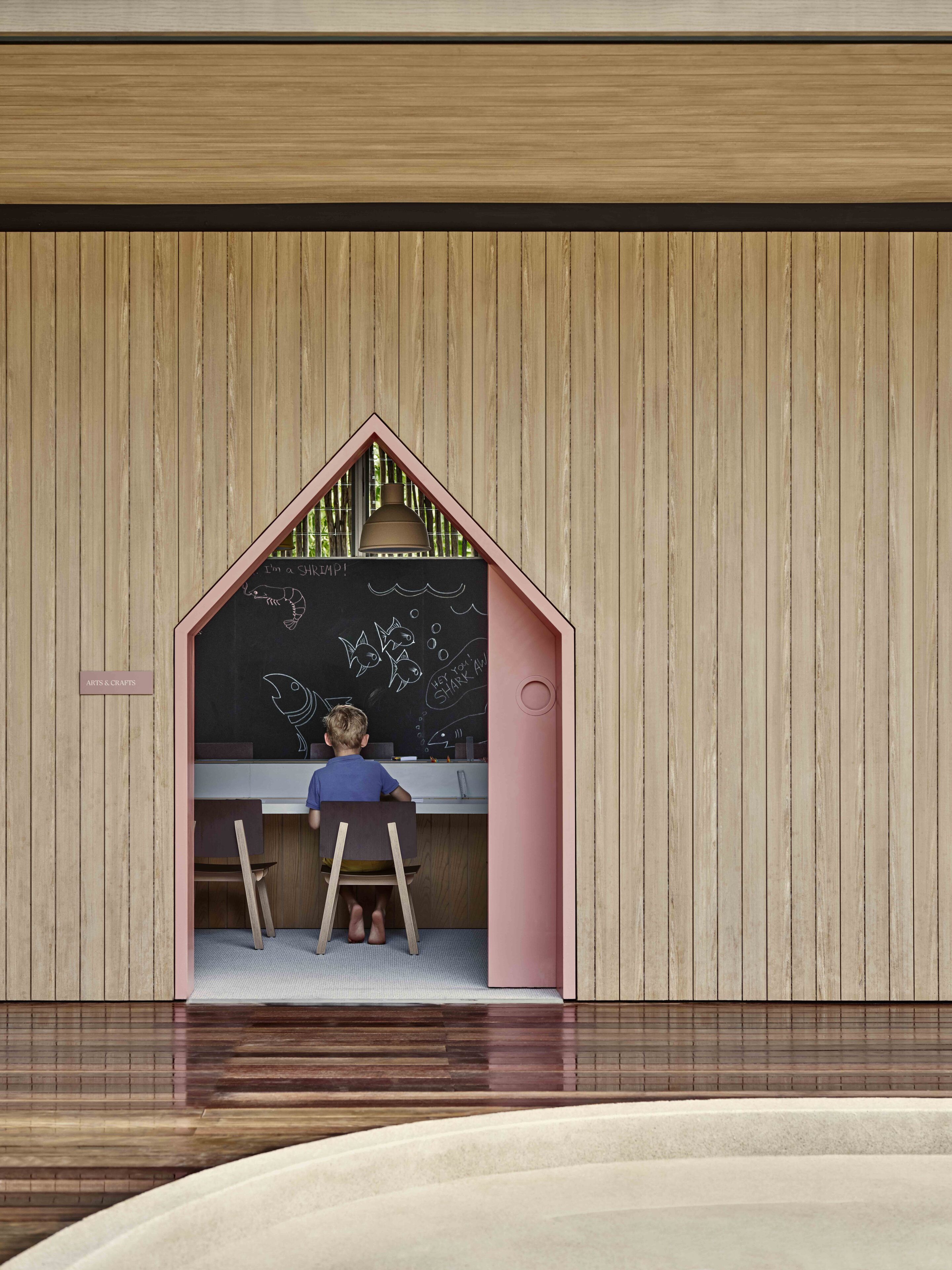 Niño dibujando en una pizarra dentro de una estructura pequeña con forma de casa y fachada de madera.