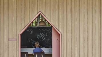Niño dibujando en una pizarra dentro de una estructura pequeña con forma de casa y fachada de madera.
