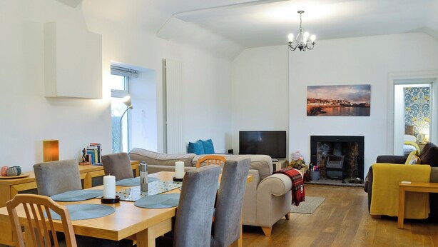 A spacious living room with a fireplace, a dining area with a wooden table and chairs, and a television. - Kirklauchline Cottage (Stoneykirk, near Portpatrick)