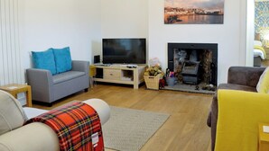 A living room with a grey sofa, a television, a fireplace, and a painting of a cityscape. - Kirklauchline Cottage (Stoneykirk, near Portpatrick)