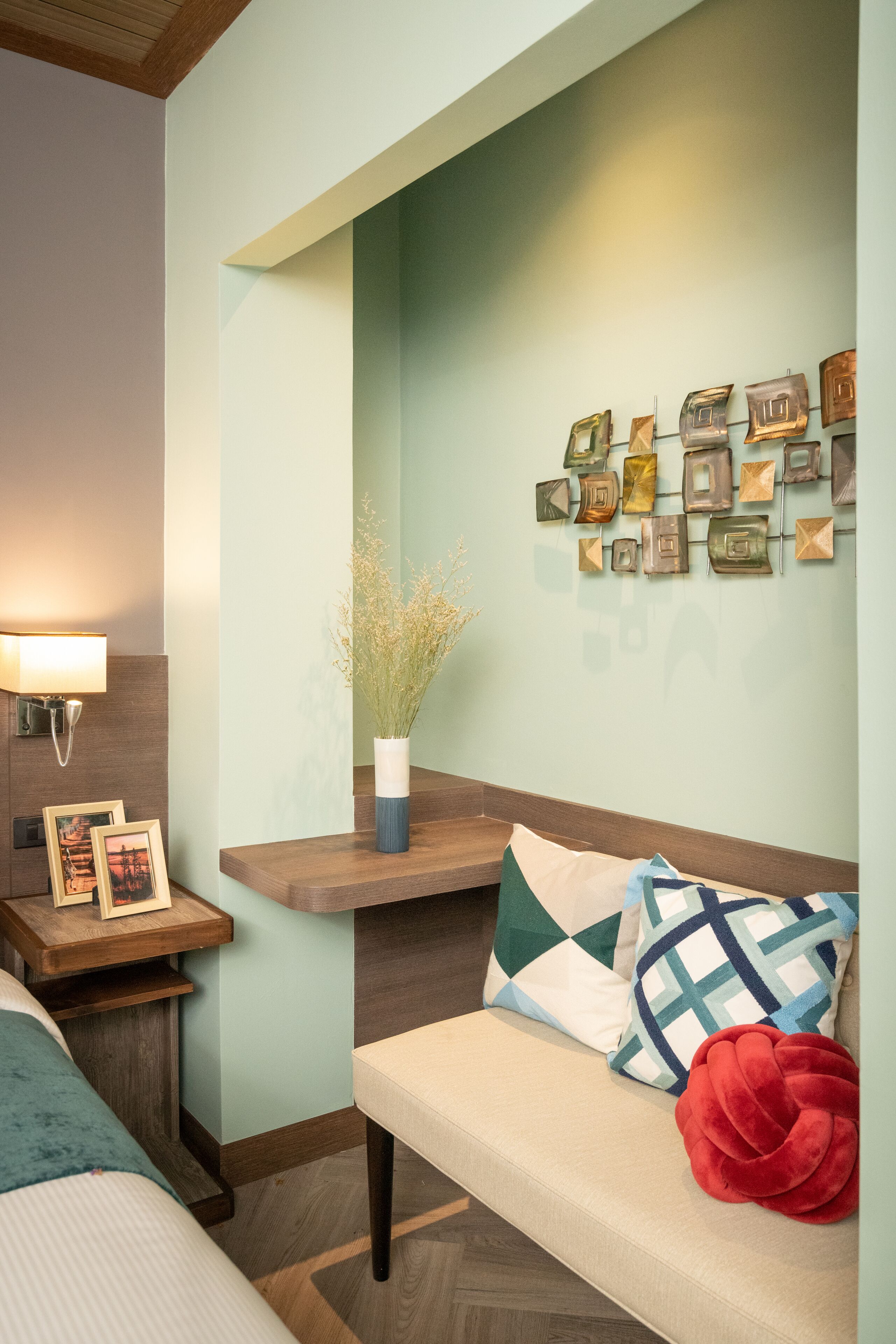 Ein Hotelzimmer mit einem Holzkopfteil, einem Nachttisch mit Lampe, einer Bank mit Kissen und einer Wand mit gerahmten Bildern.