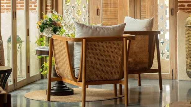 Une pièce agrémentée d’un lustre, de fauteuils en osier, d’une table ornée d’un vase de fleurs et offrant une vue sur la verdure grâce à de grandes fenêtres.
