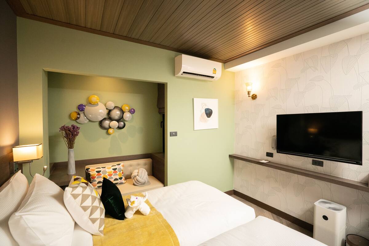 A hotel room with a bed, a TV, an air conditioner, a wall-mounted shelf, and a wall with a leaf pattern.