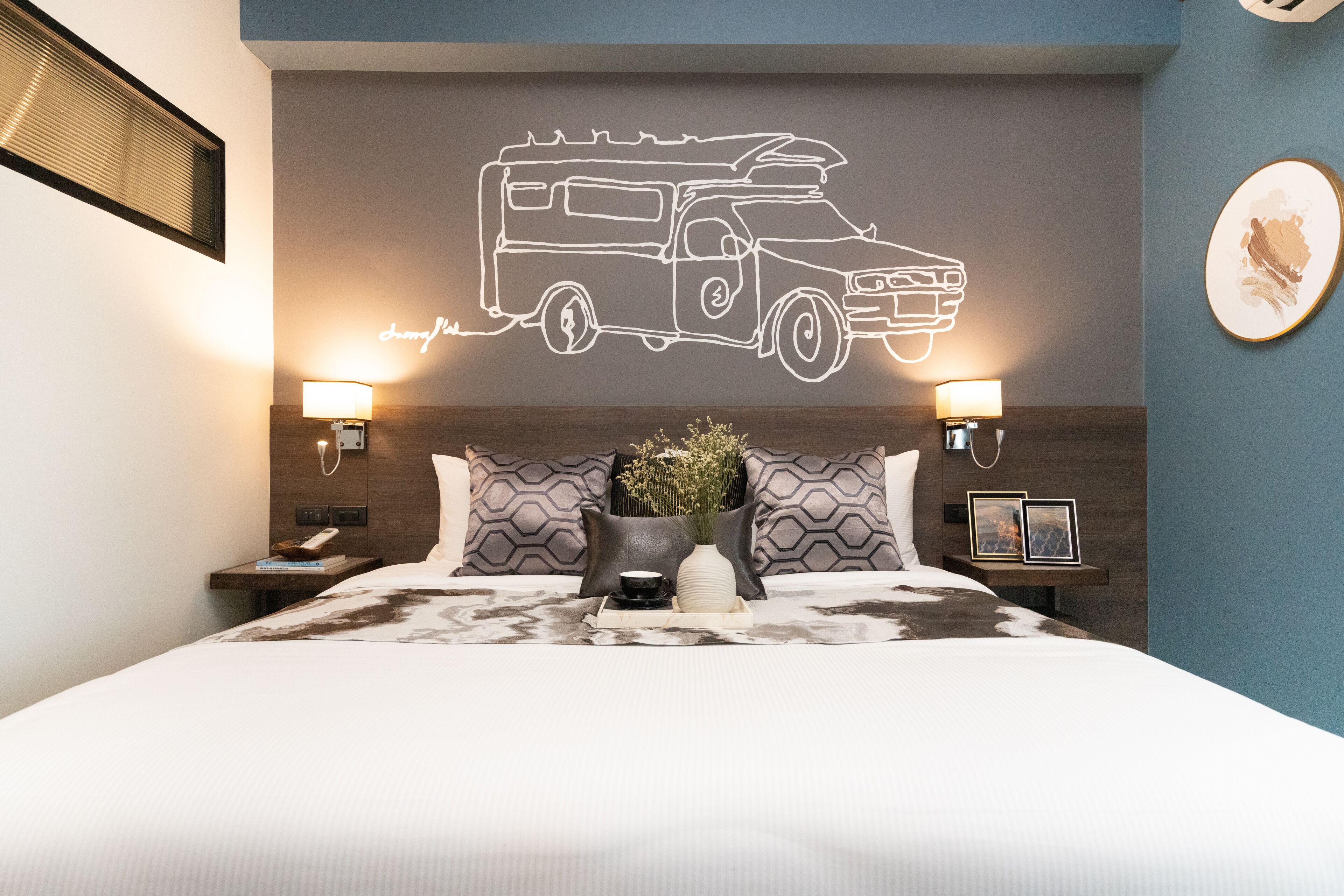 Ein Hotelzimmer mit einem Bett, Nachttischen, einer Wandleuchte, einem Wandbild mit einem Lkw und einem gerahmten Foto an der Wand.