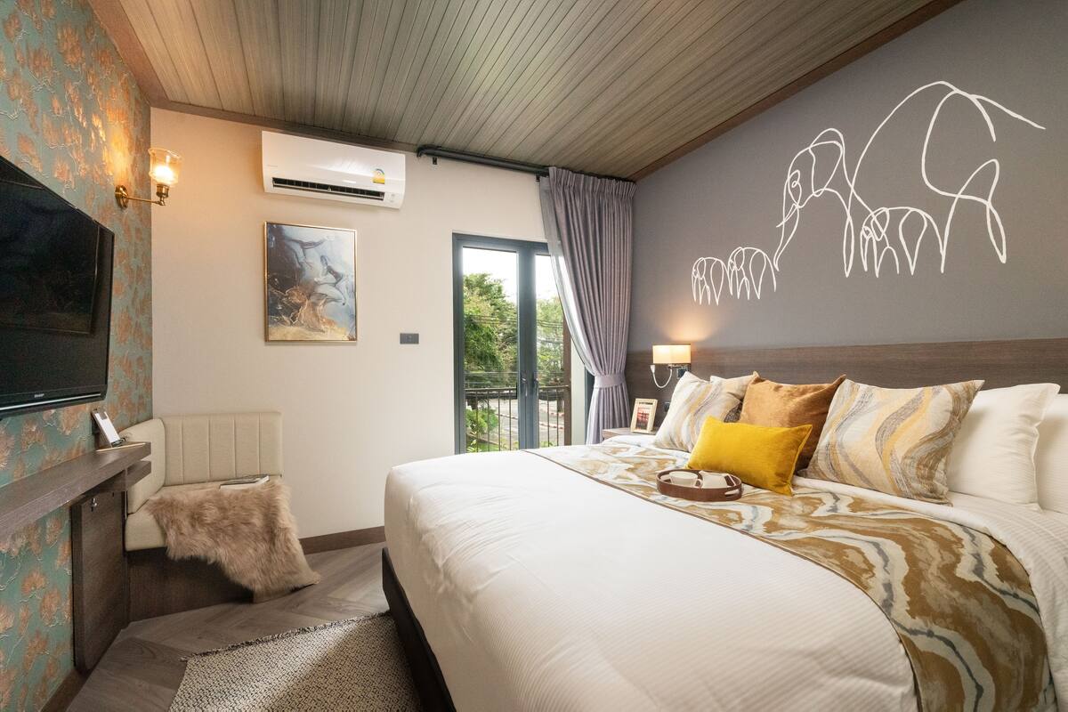A hotel room with a bed, a TV, an air conditioner, a painting, and a view of the outdoors.