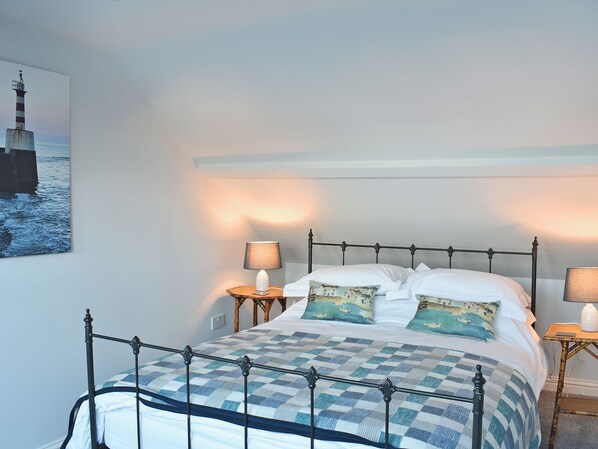 A neatly made bed with a checkered bedspread, two matching pillows, and a metal headboard. There are two bedside tables with lamps, and a framed picture of a lighthouse on the wall.