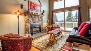 A cozy living room with a fireplace, a sofa, a patterned armchair, and a large window offering a view of trees. - Rock Pond Condo 79 | Saddleback, Ski-in/out (Sandy River Plantation)