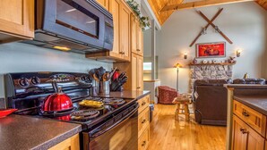 A kitchen with wooden cabinets, a stove, and a microwave. There is a stone fireplace with a mounted picture above it. - Rock Pond Condo 79 | Saddleback, Ski-in/out (Sandy River Plantation)