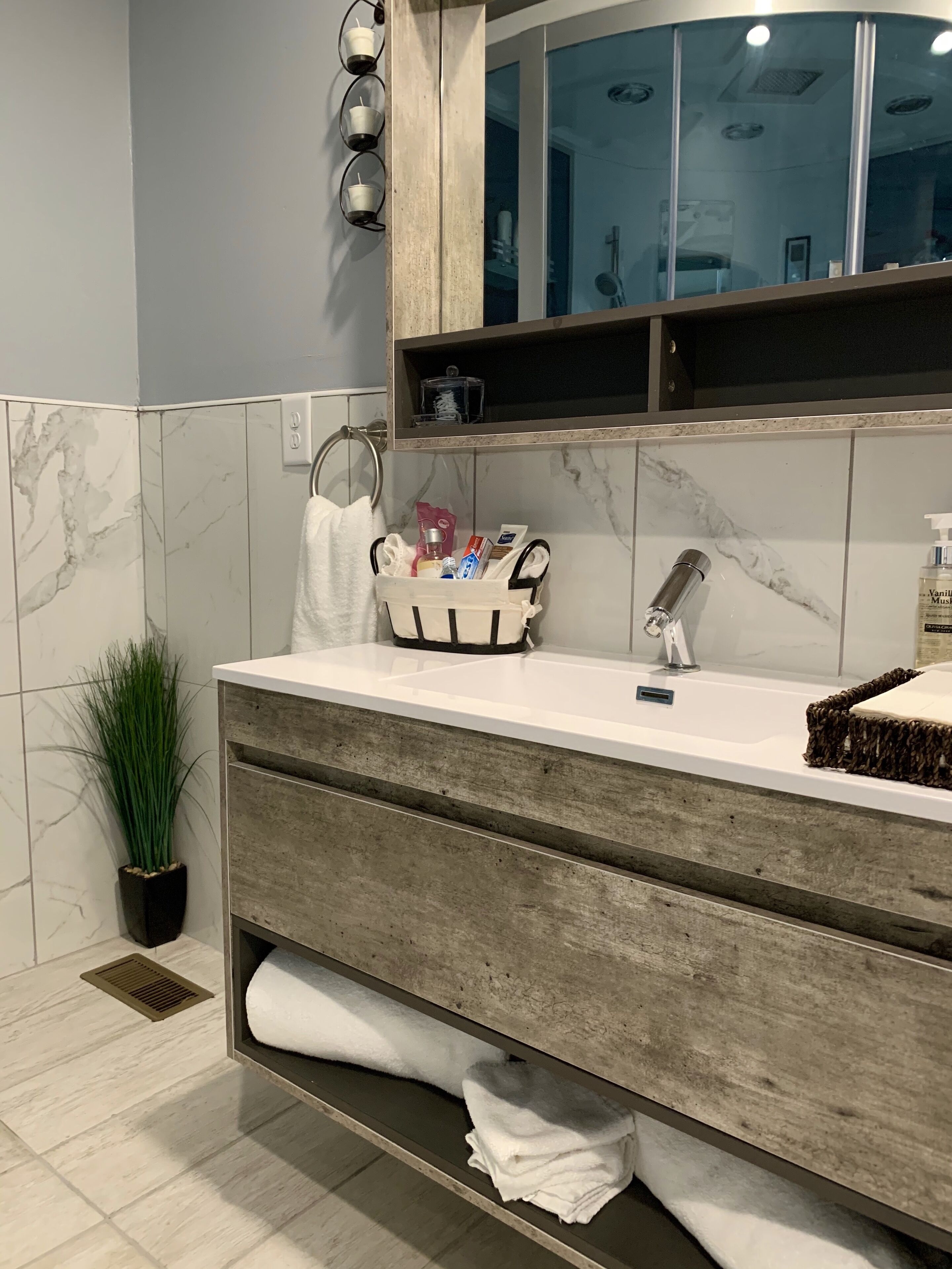 A modern bathroom with a marble-tiled wall, a wooden vanity, a glass shower enclosure, and a potted plant.