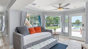 Ein geräumiges Wohnzimmer mit grauem Sofa, Deckenventilator und großen Fenstern mit Blick ins Freie.