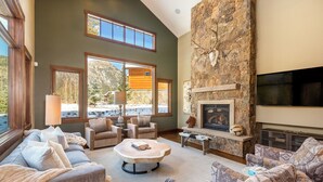 A spacious living room with a stone fireplace, comfortable seating, and large windows offering a view of a mountain cabin. - 555 Independence Road by Summit County Mountain Retreats (Keystone)