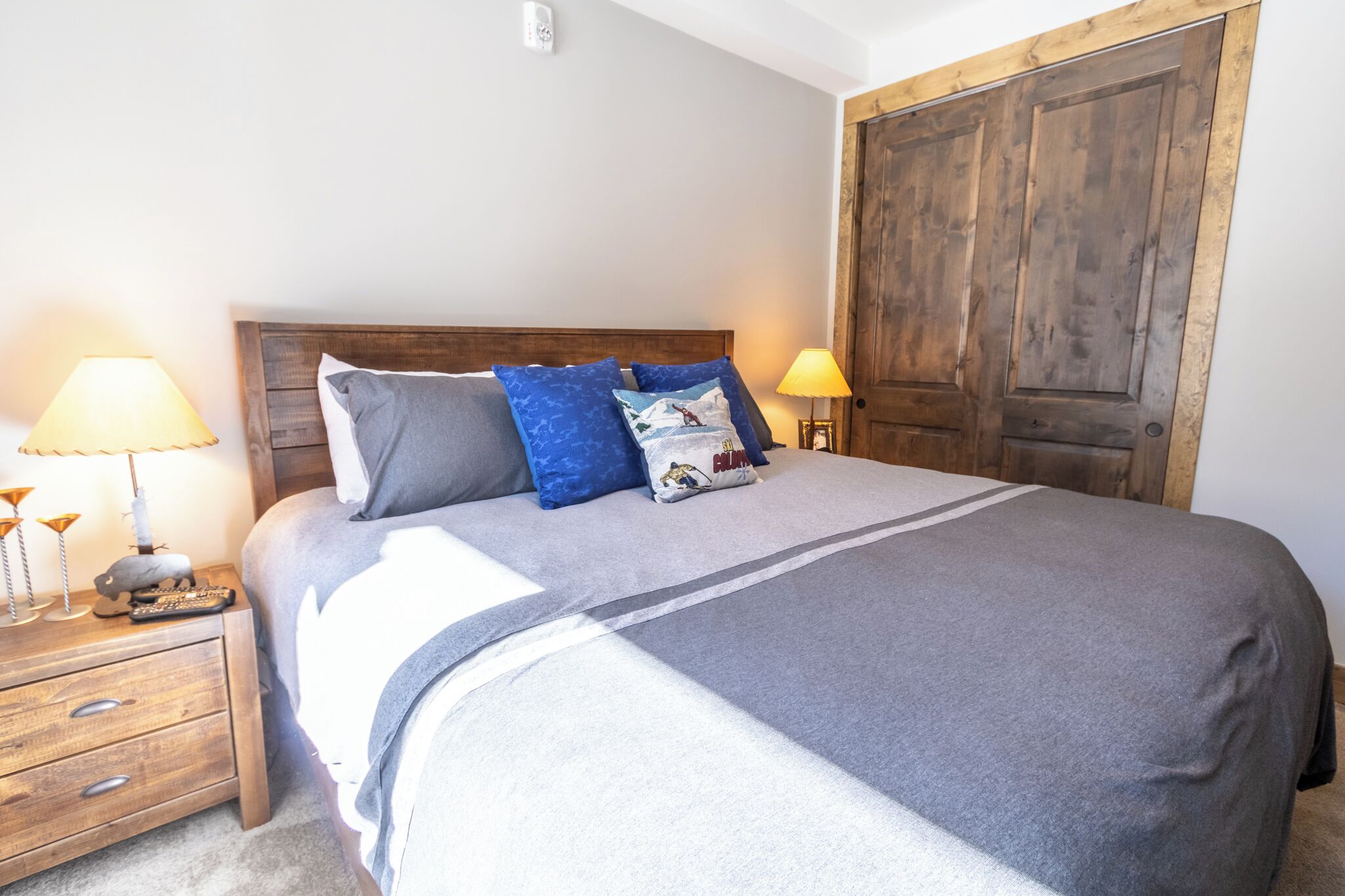 A neatly made bed with a grey and white striped bedspread, a wooden headboard, and a wooden door in the background.