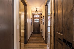 Un pasillo de madera con una puerta central, flanqueado por puertas de madera a ambos lados, que conduce a una habitación con ventana.