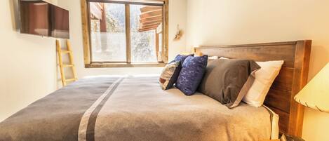 A bedroom with a large bed, a TV mounted on the wall, a window with a view, and a wooden ladder.
