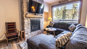 A cozy living room with a stone fireplace, a flat-screen TV mounted above it, a large window with a view of trees, a comfortable sofa, and a wooden chair. - The Seasons at Keystone #1819 by Summit County Mountain Retreats (Keystone)