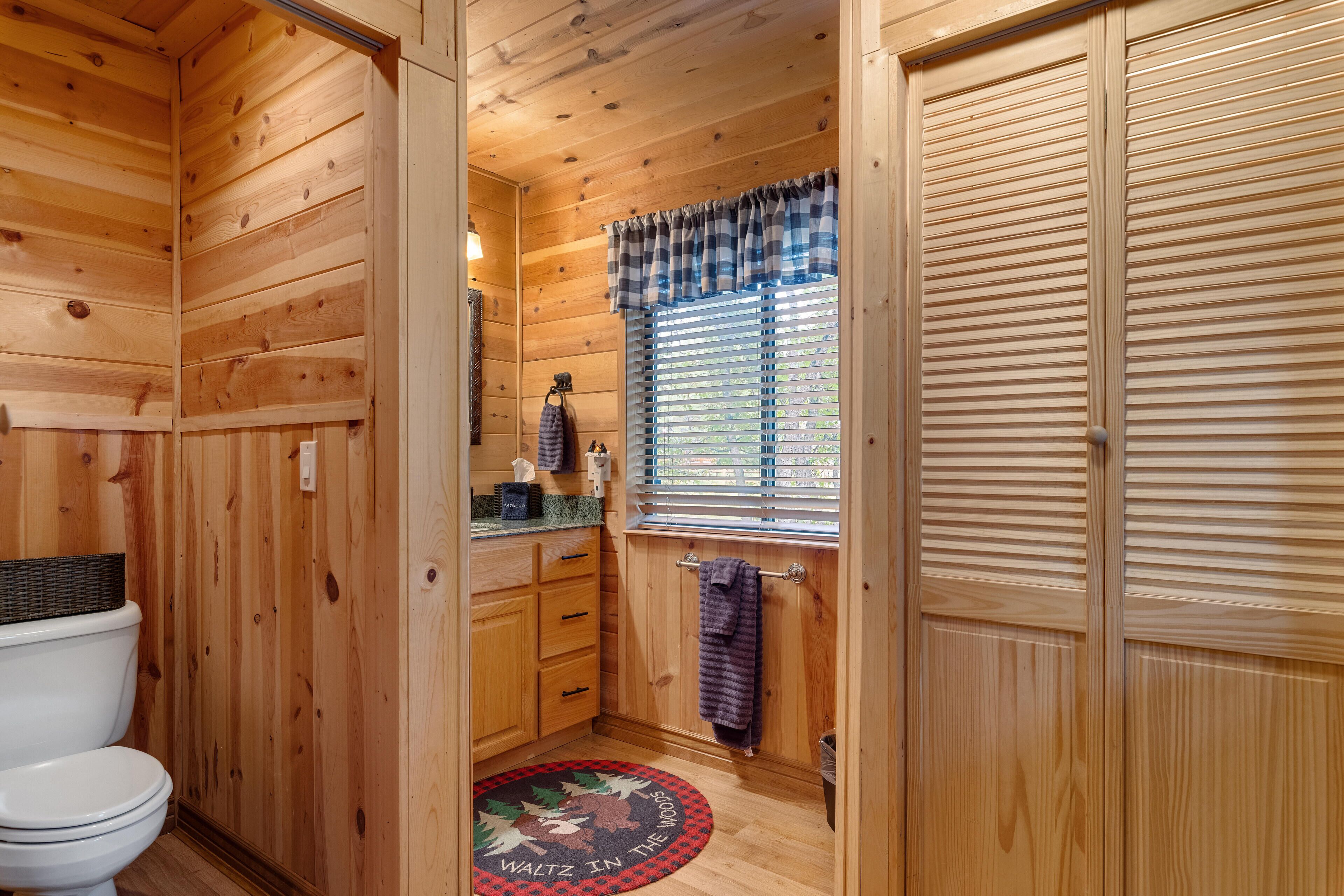 Une salle de bain rustique, aménagée dans une cabane en bois, avec des toilettes, un lavabo et une fenêtre équipée de stores.